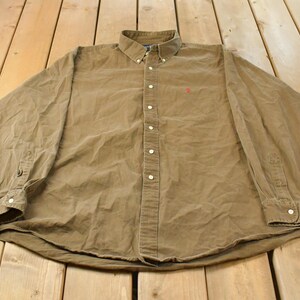 May include: A long-sleeve button-down shirt in a solid tan colour. The shirt features a button-down collar, a front button placket with white buttons, and a small red embroidered logo on the left chest. The cuffs also feature white buttons.
