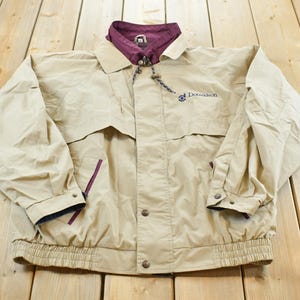 May include: Tan jacket with a burgundy collar and lining. The jacket has a button-up front, two pockets, and the word "Donaldson" embroidered on the chest. The jacket has a drawstring at the collar and a ribbed hem.
