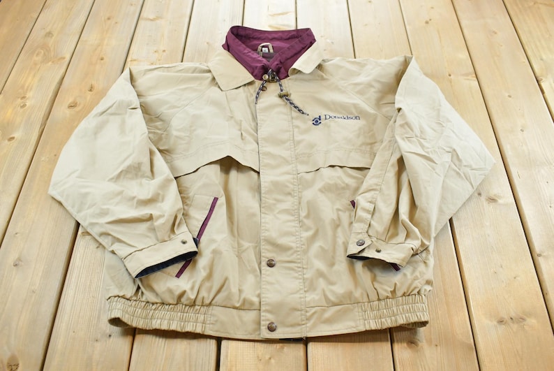 May include: Tan jacket with a burgundy collar and lining. The jacket has a button-up front, two pockets, and the word "Donaldson" embroidered on the chest. The jacket has a drawstring at the collar and a ribbed hem.
