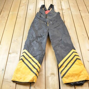 May include: Black ski overalls with yellow and black stripes down the legs. The overalls have suspenders and a red patch on the front.