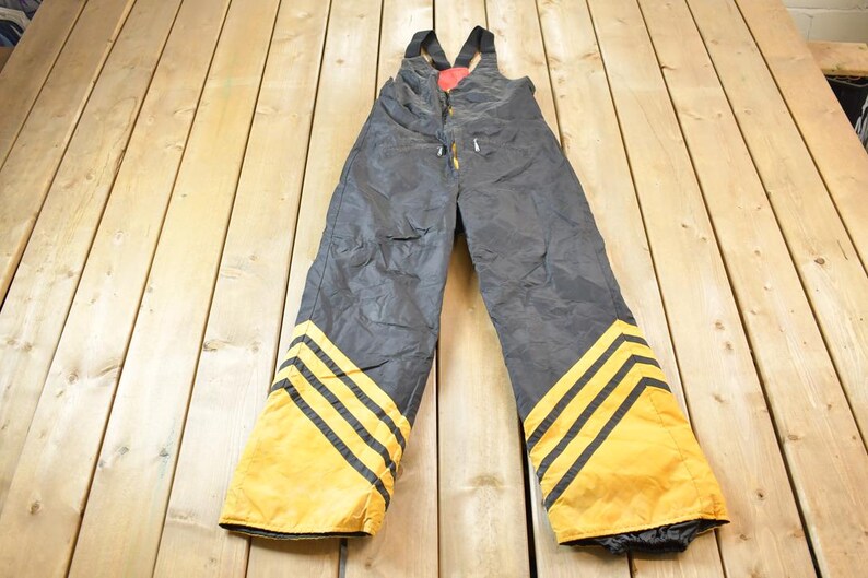May include: Black ski overalls with yellow and black stripes down the legs. The overalls have suspenders and a red patch on the front.