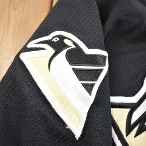 Puede incluir: Camiseta de hockey negra con un logotipo de ping&uuml;ino blanco y dorado en el hombro izquierdo. El logotipo presenta un ping&uuml;ino estilizado con un pico blanco y un cuerpo negro. La camiseta tiene un tejido de malla.