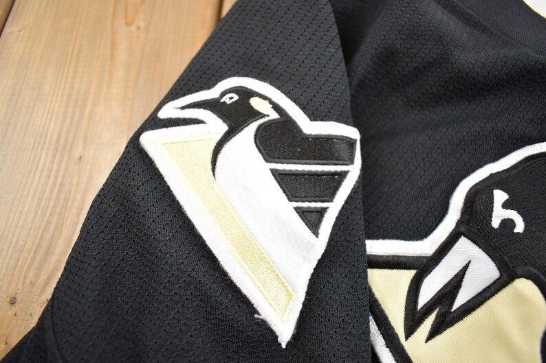 Puede incluir: Camiseta de hockey negra con un logotipo de ping&uuml;ino blanco y dorado en el hombro izquierdo. El logotipo presenta un ping&uuml;ino estilizado con un pico blanco y un cuerpo negro. La camiseta tiene un tejido de malla.