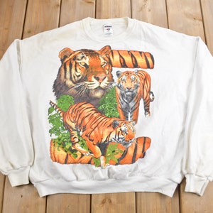 K&ouml;nnte beinhalten: Wei&szlig;er Langarm-Pullover mit einem Aufdruck von drei Tigern in einer Dschungelkulisse. Die Tiger sind orange und schwarz mit wei&szlig;en Streifen.
