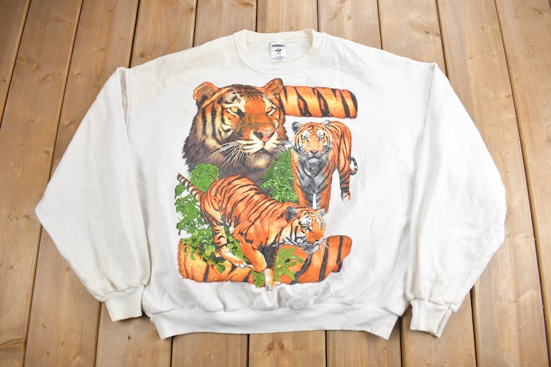 K&ouml;nnte beinhalten: Wei&szlig;er Langarm-Pullover mit einem Aufdruck von drei Tigern in einer Dschungelkulisse. Die Tiger sind orange und schwarz mit wei&szlig;en Streifen.