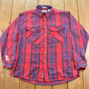 May include: A long-sleeve button-down shirt featuring a vibrant red and blue plaid pattern. The shirt includes a standard collar, a full-length front button closure, and two chest pockets with buttoned flaps. There is a visible white stain on the left sleeve.