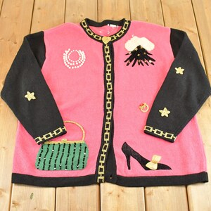 May include: A pink knit cardigan with black sleeves and a black trim featuring a gold chain pattern. The front is decorated with embroidered motifs including a green handbag, a black high-heeled shoe, stars, a pearl necklace, and a heart clasp.