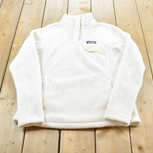 May include: A white fleece pullover with a high collar, a partial snap-button placket, and a single chest pocket featuring a Patagonia brand label. The garment is made of a textured, fuzzy material with long sleeves and a straight hemline.