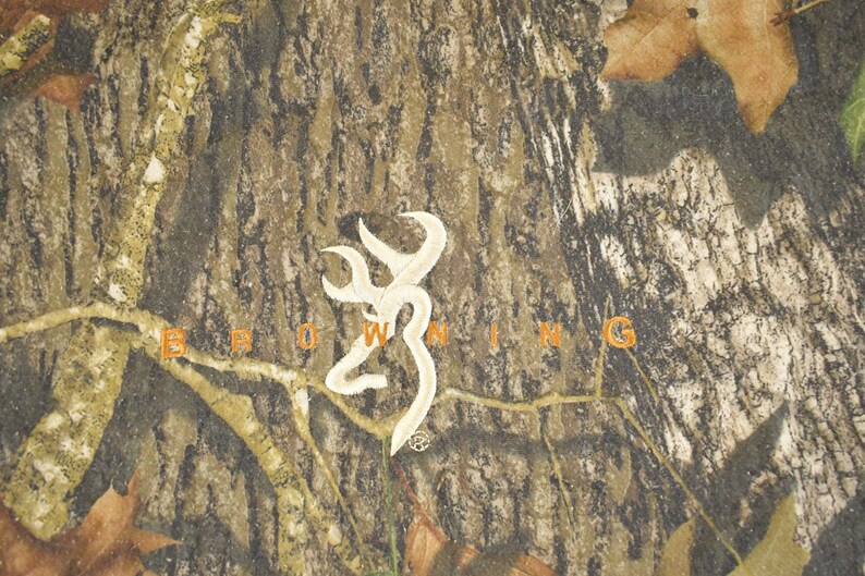 May include: Camouflage pattern with the word "BROWNING" in orange letters. A white emblem is centered above the word. The background has a mix of brown, green, and tan colours, with some leaves.