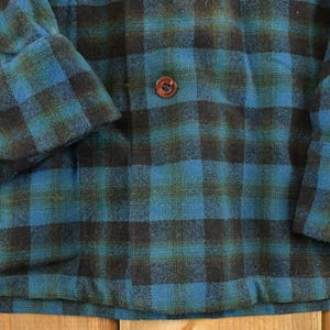 Op de afbeelding: Close-up van een blauw, zwart en groen geruit button-down shirt. Het shirt heeft een klassiek ruitpatroon met een knoopsluiting en manchetten met knopen. De stof lijkt een zacht, geweven materiaal te zijn.
