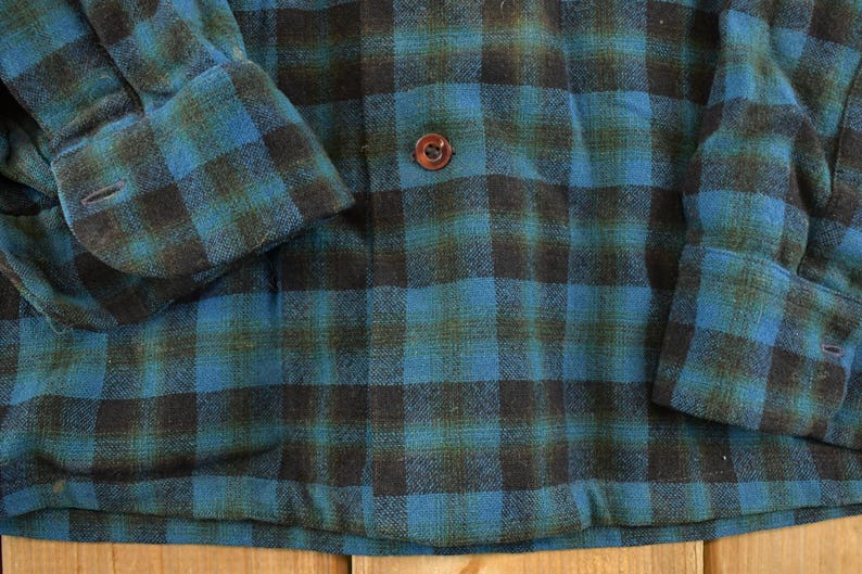 Op de afbeelding: Close-up van een blauw, zwart en groen geruit button-down shirt. Het shirt heeft een klassiek ruitpatroon met een knoopsluiting en manchetten met knopen. De stof lijkt een zacht, geweven materiaal te zijn.