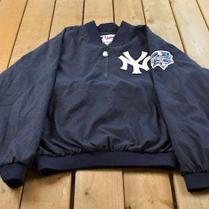 Può includere: Una giacca pullover blu navy con il logo bianco dei New York Yankees sul petto, una chiusura a zip parziale sul collo e una toppa delle World Series sulla manica sinistra. La giacca presenta maniche lunghe con polsini elastici e orlo a coste.