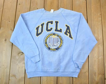 May include: A light blue sweatshirt with a large UCLA logo in navy blue and yellow. The logo includes the words "University of California" and "UCLA" in a circular design.