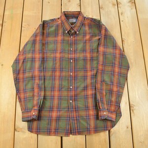 May include: A long-sleeved button-down shirt with a plaid pattern in shades of green, orange, and blue. The shirt has a button-down collar and buttoned cuffs. The garment is laid flat on a wooden surface.
