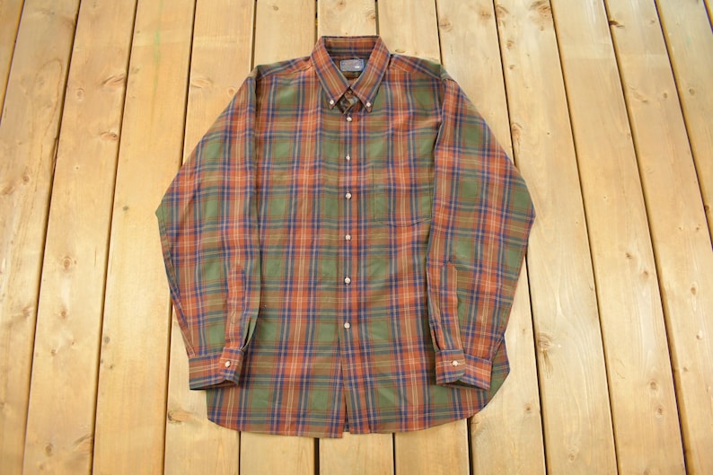 May include: A long-sleeved button-down shirt with a plaid pattern in shades of green, orange, and blue. The shirt has a button-down collar and buttoned cuffs. The garment is laid flat on a wooden surface.