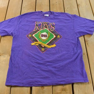 May include: Purple t-shirt with a baseball-themed graphic. The graphic features the word "KEYS" above a diamond shape with a baseball and the text "1994" inside. Below the diamond is a banner that reads "Class A Carolina League".