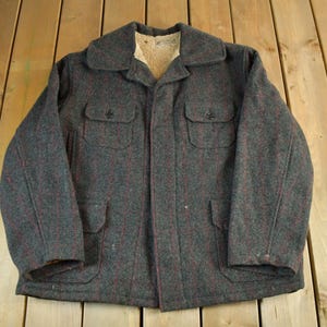 Puede incluir: Una chaqueta vintage de lana gris con un patrón moteado rojo. La chaqueta tiene cuello, cuatro bolsillos delanteros y un forro de borreguillo crema. La chaqueta está abotonada y parece estar en buen estado.