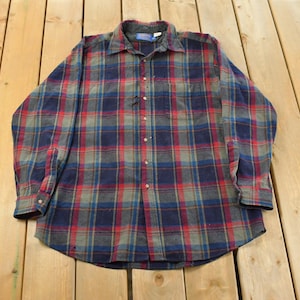 May include: A long-sleeved plaid shirt in shades of red, blue, and gray. The button-down shirt has a collared neckline and a single chest pocket. The fabric appears to be a soft flannel material, suitable for cooler weather.