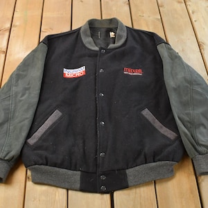 Könnte beinhalten: Schwarze und graue Vintage-Collegejacke mit grauen Ärmeln. Die Jacke hat eine Knopfleiste, zwei Vordertaschen und einen grauen gerippten Kragen, Bündchen und Saum. Mit den Logos "INGRAM MICRO" und "maxell".