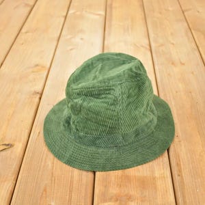 Può includere: Un cappello a secchiello verde in velluto a coste con una corona arrotondata e una tesa corta. Il cappello è di un colore verde opaco e uniforme e poggia su una superficie di legno.