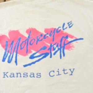 May include: Cream-colored t-shirt with a graphic design. The design features the words "Motorcycle Stuff" in blue script over a pink brushstroke, with "Kansas City" below in blue. The shirt is made of a soft fabric.