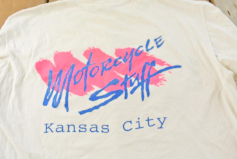 May include: Cream-colored t-shirt with a graphic design. The design features the words "Motorcycle Stuff" in blue script over a pink brushstroke, with "Kansas City" below in blue. The shirt is made of a soft fabric.