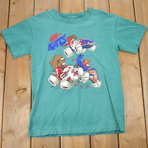 Peut inclure: Un t-shirt bleu turquoise avec un motif de trois animaux de dessin animé conduisant des karts. Le texte "40-KATS" est imprimé en lettres bleues et rouges sur le t-shirt.