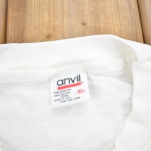 Puede incluir: Camiseta blanca con una etiqueta blanca que dice "Anvil" y "XL" en letras rojas.