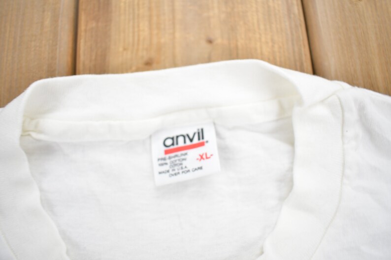 Puede incluir: Camiseta blanca con una etiqueta blanca que dice "Anvil" y "XL" en letras rojas.
