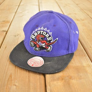 Puede incluir: Gorra de béisbol morada y negra de los Raptors. La gorra presenta el logotipo de los Raptors, un dinosaurio rojo sosteniendo un balón de baloncesto. Tiene una visera plana negra y una corona morada. La gorra está sobre una superficie de madera.