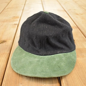Puede incluir: Una gorra negra con una visera de ante verde. La gorra tiene una corona redondeada y una visera plana. La gorra está hecha de un material similar a la lana. La visera es de color verde claro. La gorra está sobre una superficie de madera.