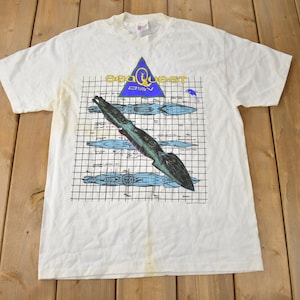 May include: White t-shirt featuring a graphic print of a submarine and the text "Seaquest DSV".