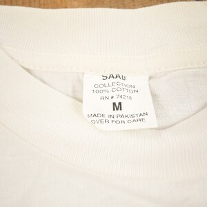 May include: White cotton t-shirt tag with black text that reads "SAAL COLLECTION 100% COTTON RN # 74218 M MADE IN PAKISTAN OVER FOR CARE"