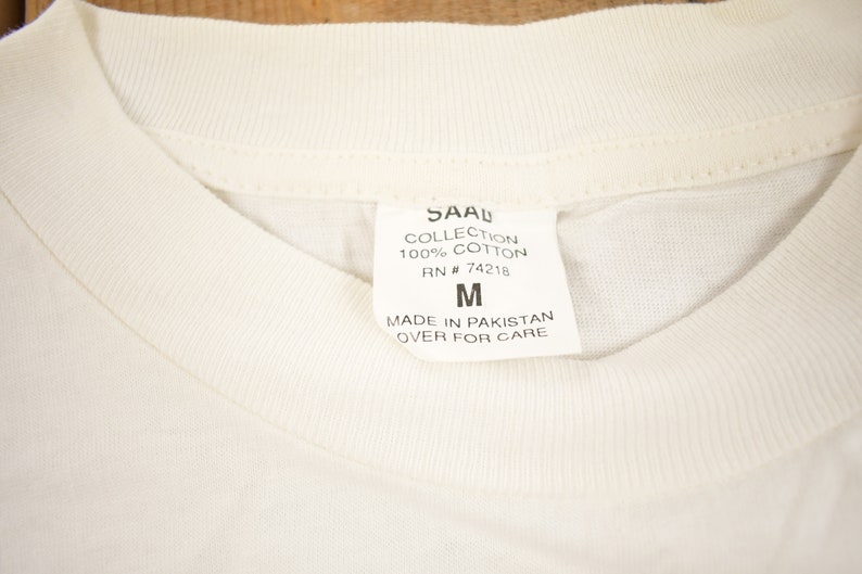 May include: White cotton t-shirt tag with black text that reads "SAAL COLLECTION 100% COTTON RN # 74218 M MADE IN PAKISTAN OVER FOR CARE"