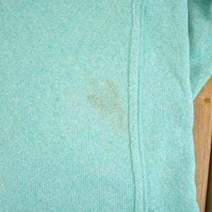 May include: Close-up of a light teal-colored knit fabric. The fabric has a textured appearance and a visible seam running vertically. A brown stain is present on the fabric.