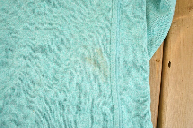 May include: Close-up of a light teal-colored knit fabric. The fabric has a textured appearance and a visible seam running vertically. A brown stain is present on the fabric.