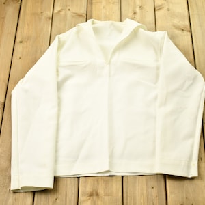Puede incluir: Una camisa blanca de manga larga con cuello marinero. La camisa está colocada sobre una superficie de madera.