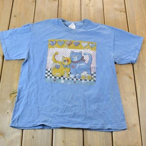 May include: A light blue t-shirt with a graphic of two cats. The cats are sitting next to each other and are surrounded by hearts and flowers. The text "Love is in the air" is printed on the shirt.
