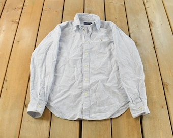 May include: A light blue and white striped button-down shirt. The long-sleeved shirt has a collar and a small logo on the chest. The shirt is made of a lightweight fabric and is laid flat on a wooden surface.