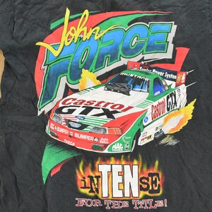 Op de afbeelding: Zwart t-shirt met een kleurrijke afbeelding van een raceauto, de tekst "John Force" en "Castrol GTX." Het ontwerp bevat vlammen en de woorden "Intense for the Title!"