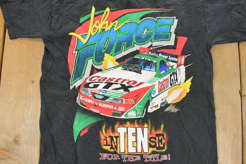 Op de afbeelding: Zwart t-shirt met een kleurrijke afbeelding van een raceauto, de tekst "John Force" en "Castrol GTX." Het ontwerp bevat vlammen en de woorden "Intense for the Title!"