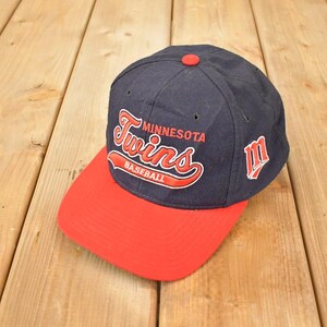 May include: A navy blue baseball cap with a red peak. The cap displays the text "Minnesota Twins Baseball" in red script. A red button is on top, and a red "M" logo is on the side.