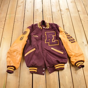 May include: A maroon varsity jacket with brown leather sleeves. The jacket has a large "E" on the front and the name "Ashley" embroidered on the left side. The sleeves have yellow and white stripes and patches with the words "Evansville Victors".