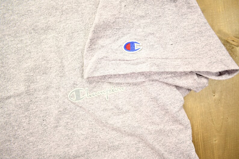 May include: A heather grey t-shirt with a white Champion logo on the right sleeve and a white Champion logo on the front of the shirt.