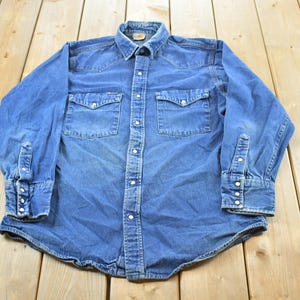 Puede incluir: Una camisa vaquera azul con dos bolsillos en el pecho y cierres de botón. La camisa de manga larga tiene cuello y puños abotonados. La camisa está tendida sobre una superficie de madera.