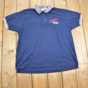 May include: A navy blue polo shirt with a grey collar and white trim on the sleeves. The shirt features the word "DARE" in red script on the chest, with additional text below. The shirt is made of a soft material.