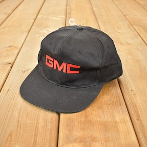 Puede incluir: Gorra de béisbol negra con las letras rojas "GMC" bordadas en la parte delantera. La gorra tiene una visera curva y una corona estructurada. El sombrero está sobre una superficie de madera.