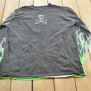 May include: Dark gray long-sleeve shirt with a skull and crossbones graphic on the back. The sleeves feature a green and purple flame design. The shirt has a green trim at the bottom.
