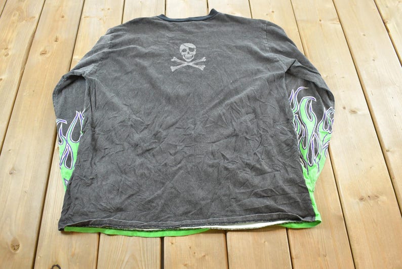 May include: Dark gray long-sleeve shirt with a skull and crossbones graphic on the back. The sleeves feature a green and purple flame design. The shirt has a green trim at the bottom.