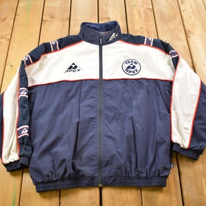 May include: A navy blue, white, and red windbreaker jacket with a zipper closure. The jacket has a white panel on the front with the logo "Team Apex" in a circle. The left side of the jacket has the logo "Apex" in white.